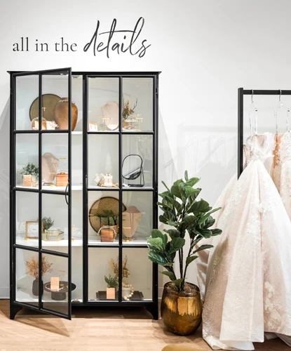 A modern bridal boutique display showcasing a sleek black cabinet filled with wedding accessories such as jewelry, hairpieces, and decorative items. To the right, a rack of luxurious white and blush wedding dresses is partially visible. A gold planter with a vibrant green plant adds a natural accent to the scene. The phrase "all in the details" is stylishly displayed on the wall above the cabinet, emphasizing attention to the boutique's curated offerings. The setting is well-lit, with light wood flooring contributing to a contemporary and inviting ambiance.