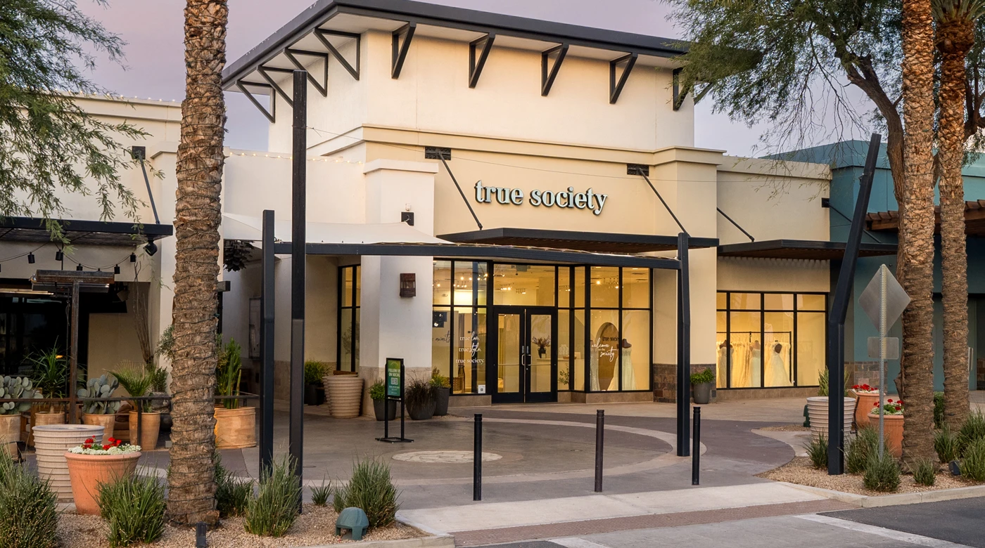 Exterior view of the True Society bridal boutique in Peoria Arizona during twilight, showcasing a modern architectural design with clean lines and a neutral color palette. The boutique features large, illuminated display windows showcasing elegant wedding dresses. The entrance is framed by stylish black metal accents and surrounded by desert landscaping, including palm trees, cacti, and planters with vibrant flowers. Soft string lights add warmth to the inviting storefront, emphasizing its sophisticated ambiance.