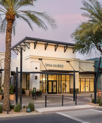 An exterior view of the True Society bridal boutique in Peoria Arizona at twilight, featuring a modern design with illuminated display windows showcasing wedding dresses. The entrance is highlighted with black metal accents and surrounded by desert landscaping, including palm trees and decorative planters. The soft glow of string lights enhances the inviting ambiance of the storefront.