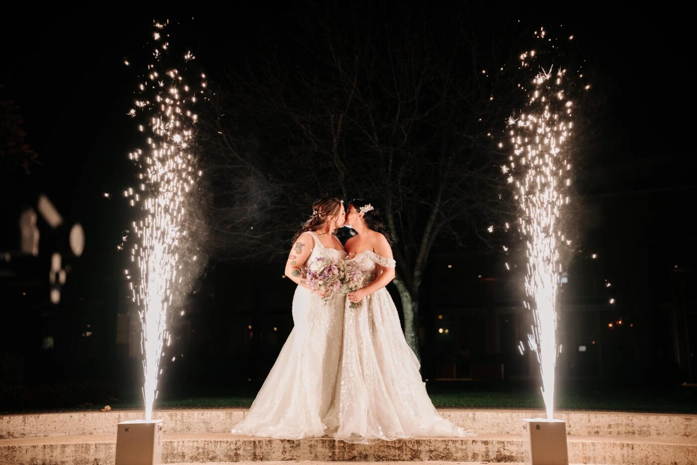 Brides wearing lace wedding dresses kissing with fireworks going off 