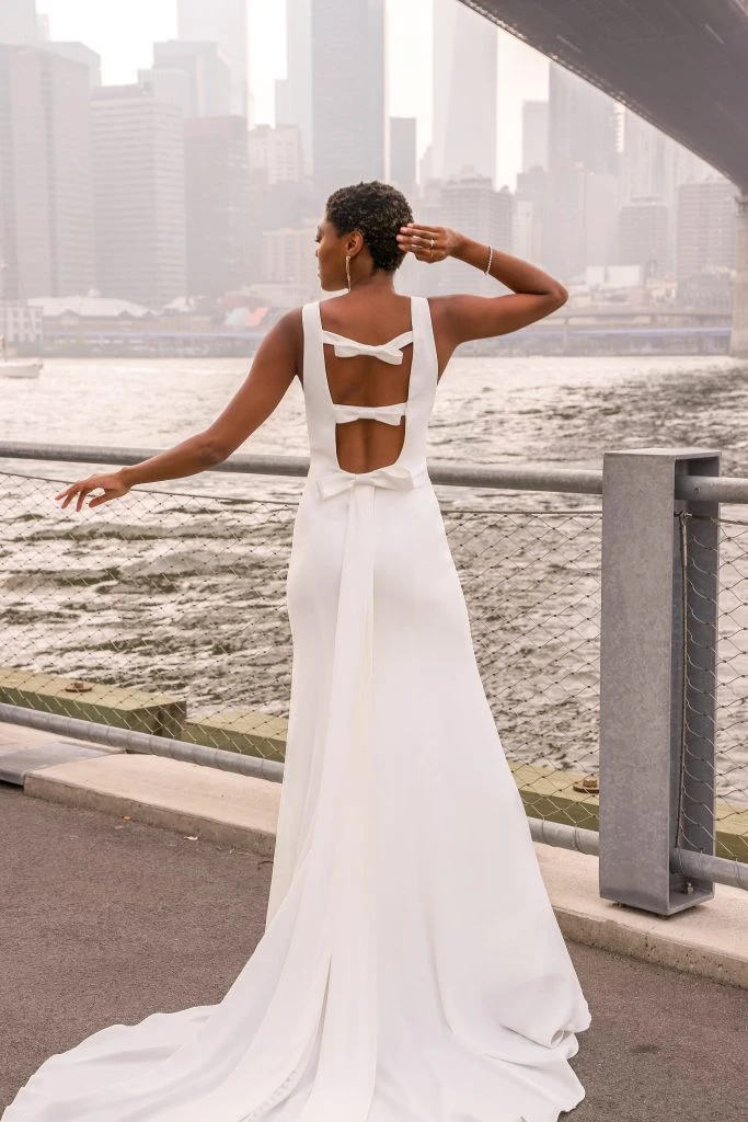 Bride facing the river with skyline in the background. She is wearing a simple high neck wedding dress with 3 bows along the back