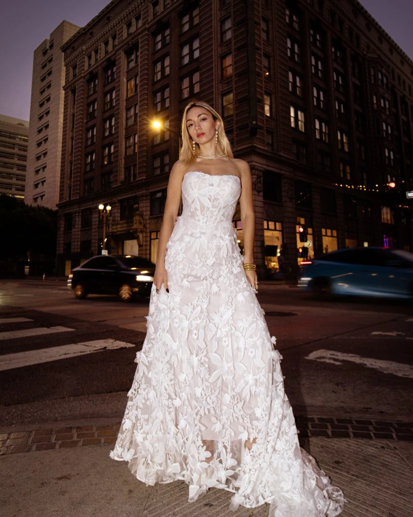 Bride standing in near a busy street wearing a strapless floral lace wedding dress