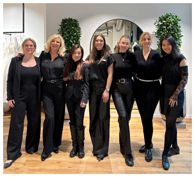 Group photo of seven True Society True Crew bridal stylists wearing all-black outfits, smiling and posing inside a modern bridal boutique with gowns in the background. The team stands on wood flooring in front of a round mirror and decorative plants, showcasing a welcoming and professional bridal shopping environment.
