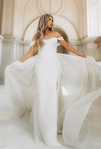 Petite bride wearing sparkling off-the-shoulder wedding dress with overskirt