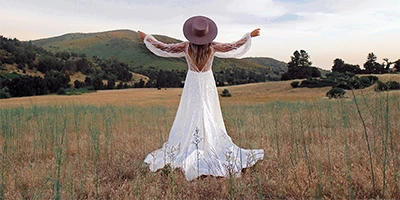 bride standing with hands out wide in a field wearing a wedding dress and brimmed hat