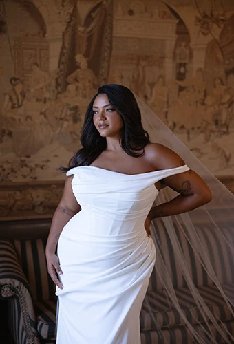 Plus-size bride wearing crepe sheath wedding gown with asymmetric off-shoulder straps and ruched corset bodice, Essense of Australia D4563+